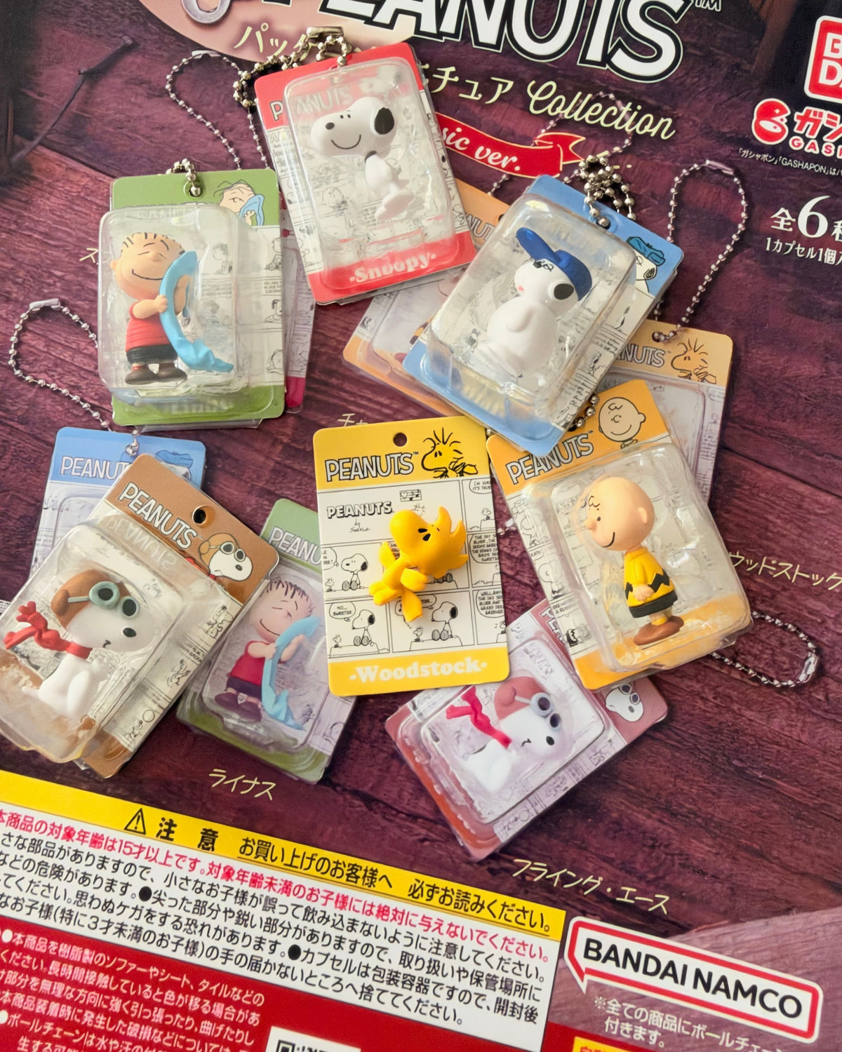 A collection of Peanuts character keychain boxes and small character figures arranged on top of a branded Peanuts display card. Visible brands include Peanuts and Bandai Namco.