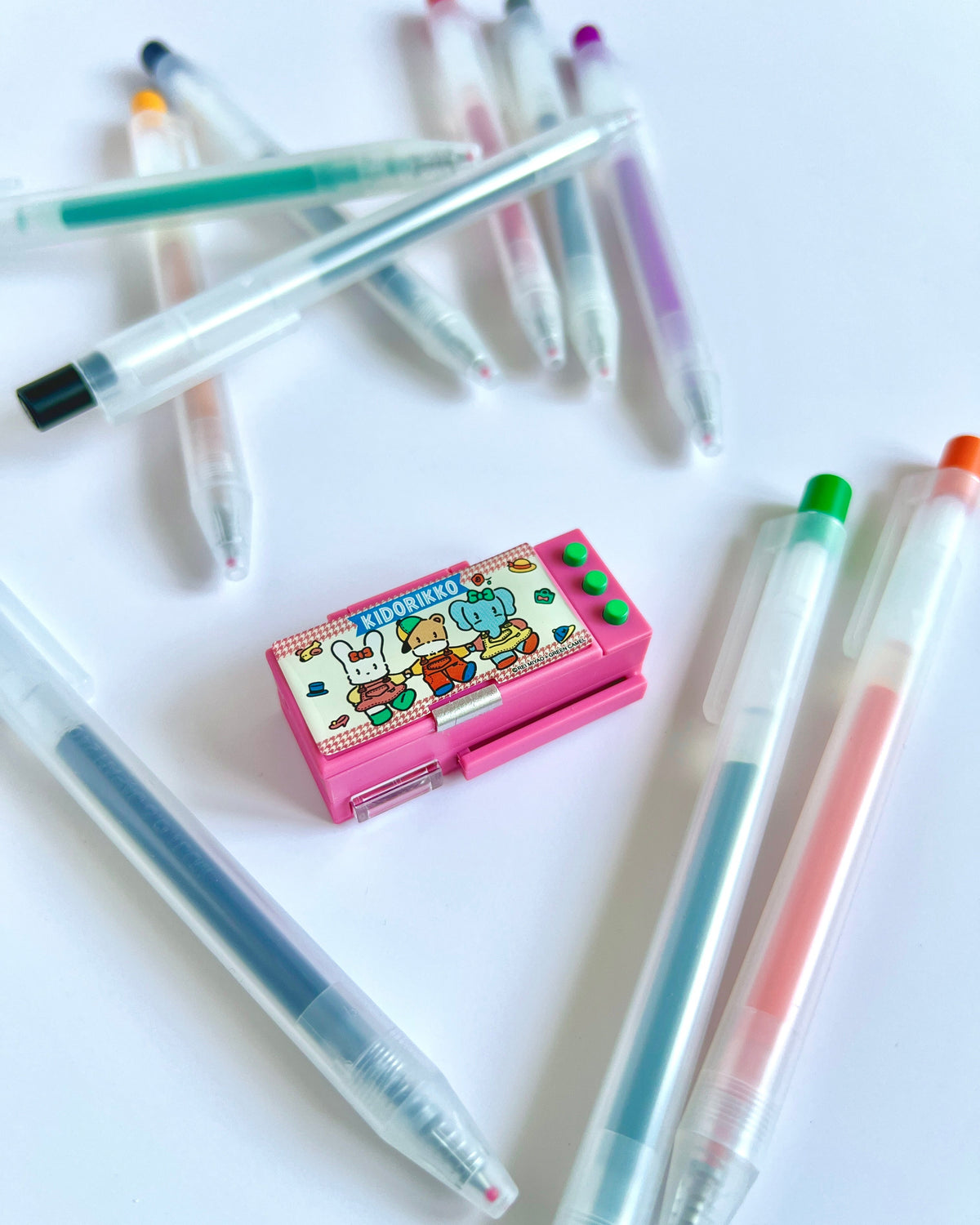 A pink cartoon-themed pen case with a rabbit illustration sits on a white surface, surrounded by several translucent gel pens in assorted colors.